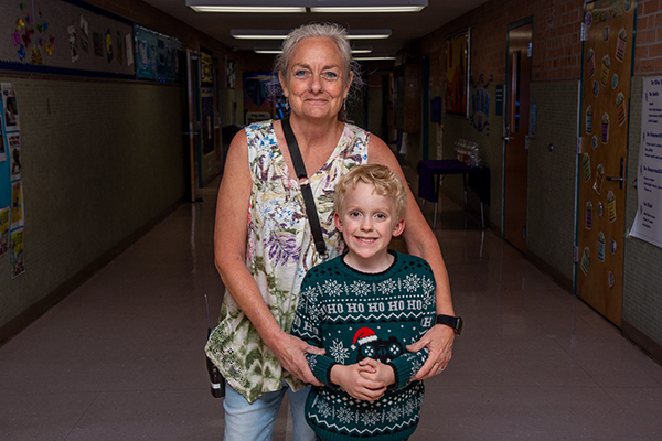 A woman in a floral shirt stands in the school hallway with her young grandson