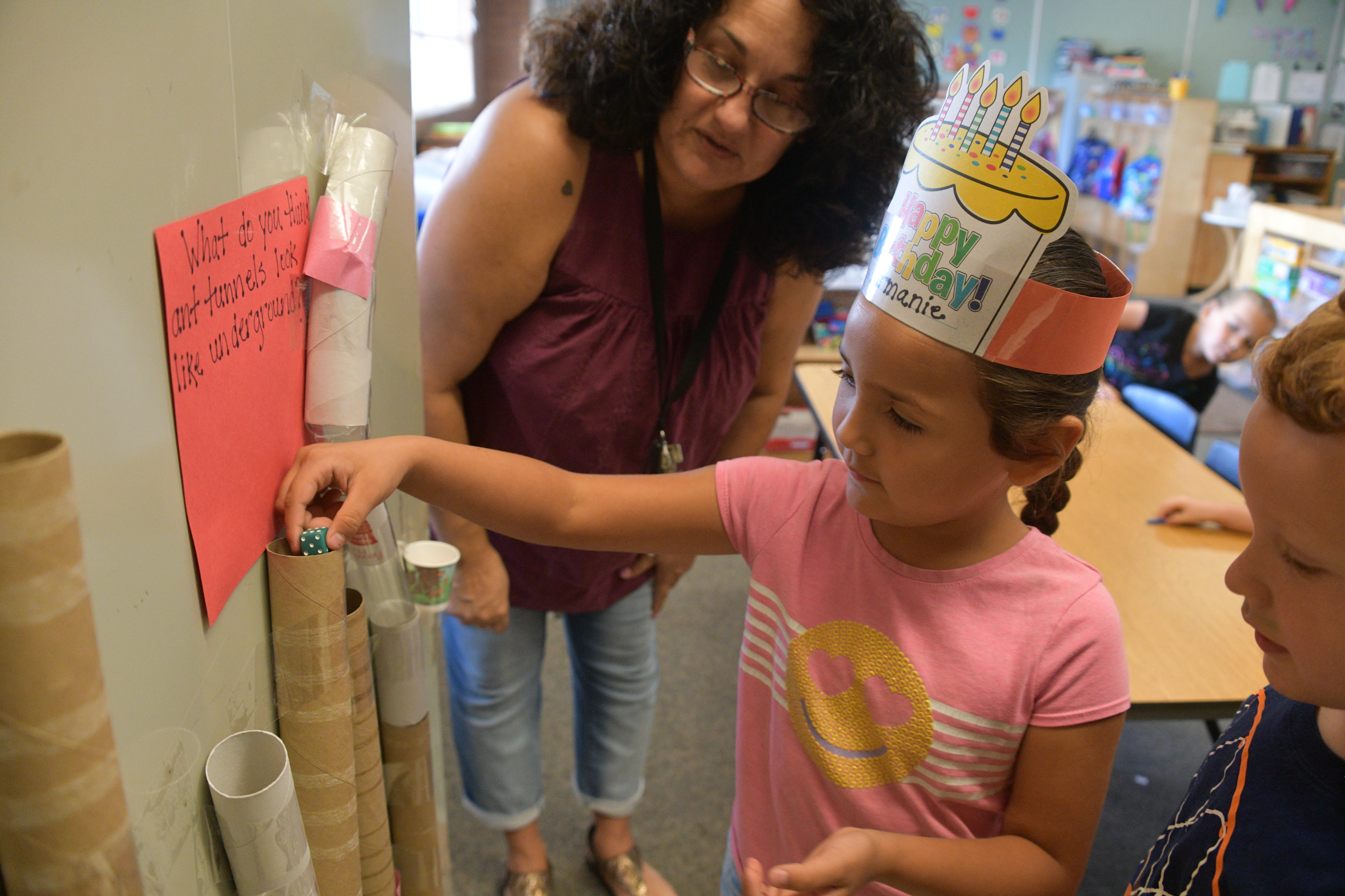 A girl in a birthday crown drops a die down a paper towel tube while her teacher and a fellow student look on